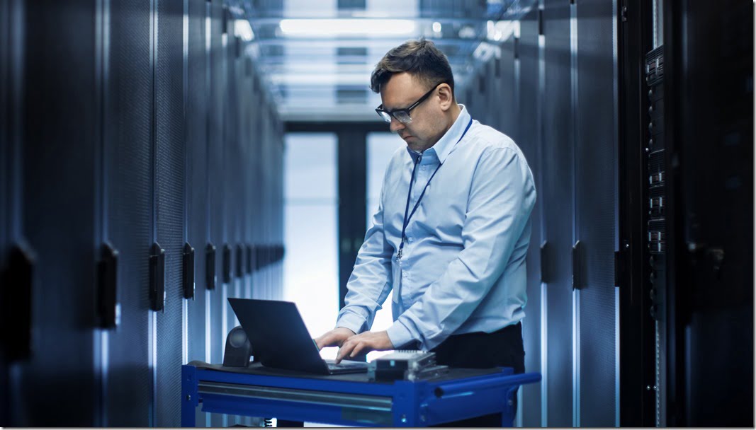 In Data Center IT Engineer Works on Crash Cart Laptop. We See Rows of Server Racks and One Cabinet is Open.